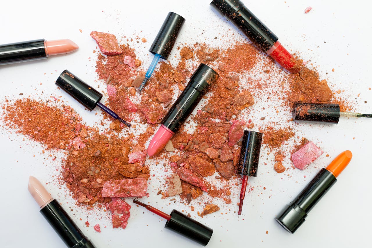 hero-img Colorful scattered makeup products including lipsticks and crushed powders on a white background.