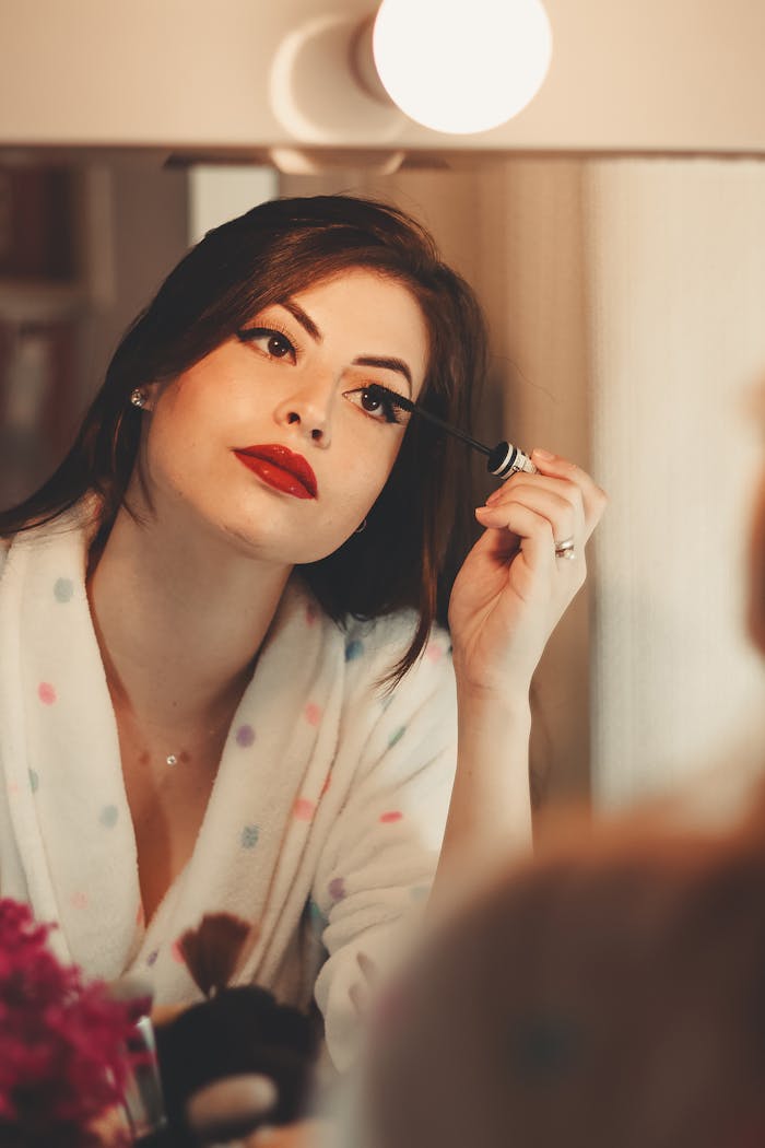 experience-img Glamorous woman applying mascara in front of a vanity mirror, enhancing her beauty.