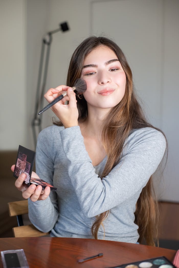 A young woman cheerfully applying makeup with a brush indoors.